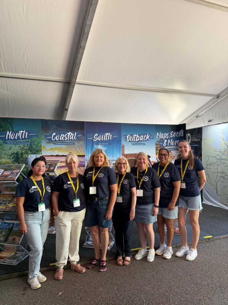 Seven people wearing matching navy shirts and yellow lanyards stand in a row inside a tent with travel-themed banners displayed behind them.