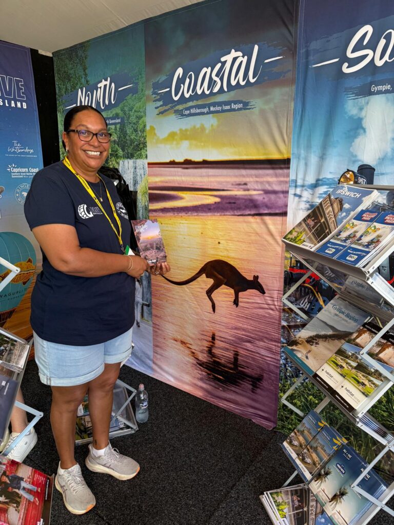 A woman stands next to a display booth featuring regional travel brochures and a large poster of a kangaroo by the water labeled “Coastal.”.