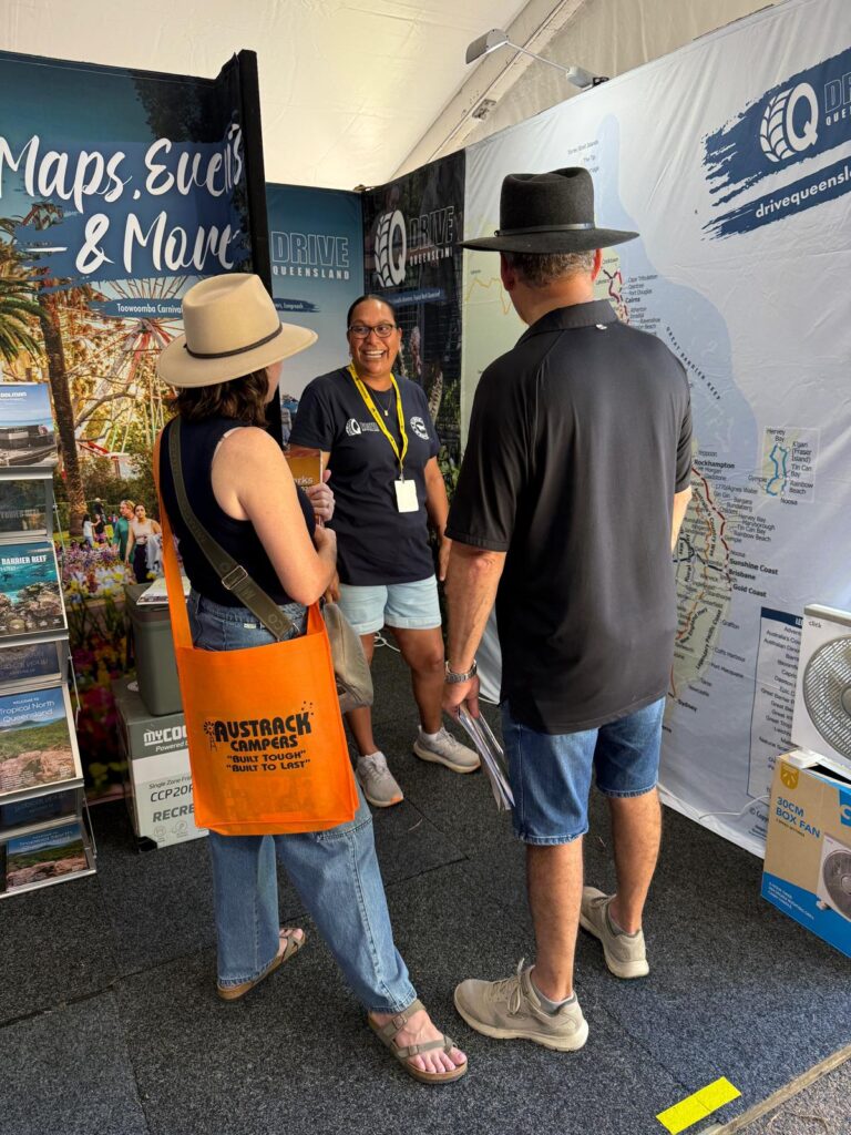 Three people stand and talk at an indoor event booth featuring tourism maps, brochures, and a large map display for Drive Queensland.