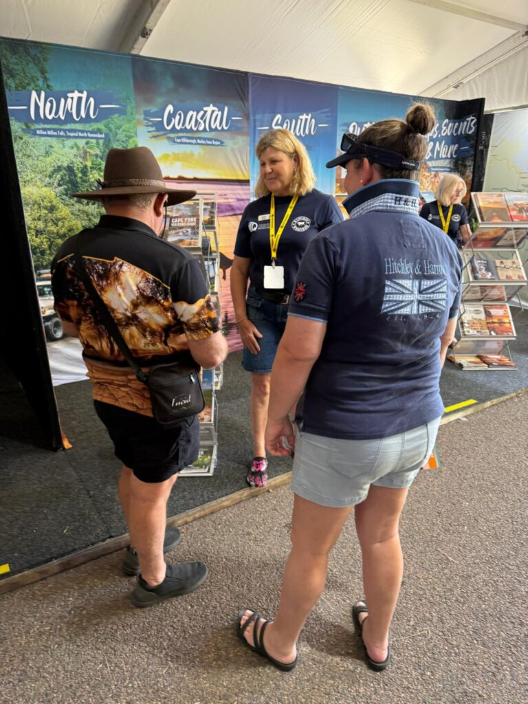 Three people stand and talk at an indoor event booth featuring travel posters and regional displays under a tent.