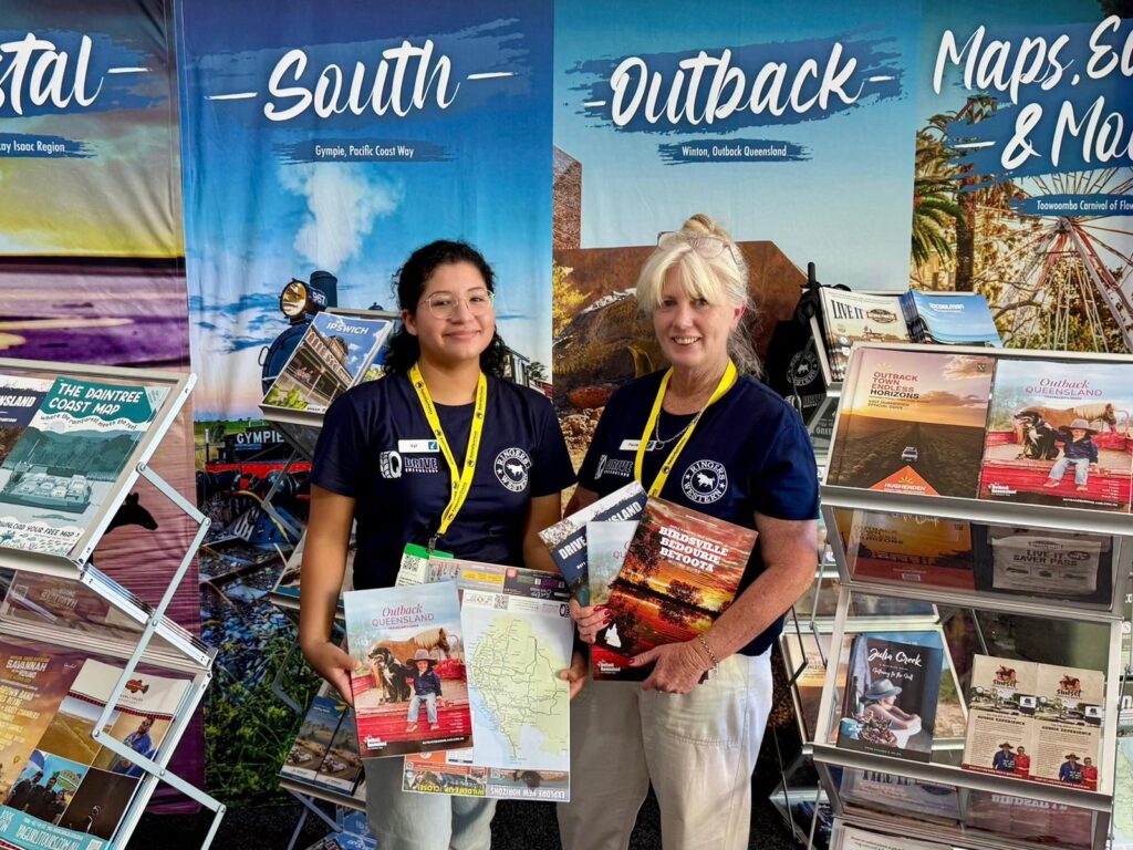 Two women wearing name tags and navy shirts stand in front of tourism brochures and maps, smiling and holding maps at a promotional booth with travel posters in the background.