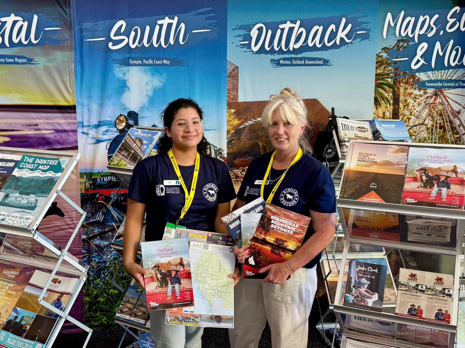 Two women wearing name tags and navy shirts stand in front of tourism brochures and maps, smiling and holding maps at a promotional booth with travel posters in the background.