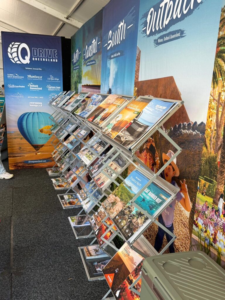 A display rack holds various tourism brochures in front of large banners promoting different regions such as Outback, Coast, and South in Queensland, Australia.