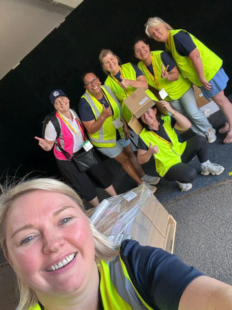 Six people wearing neon safety vests smile and pose for a group photo in front of a black backdrop, with boxes and pallets visible in the foreground.