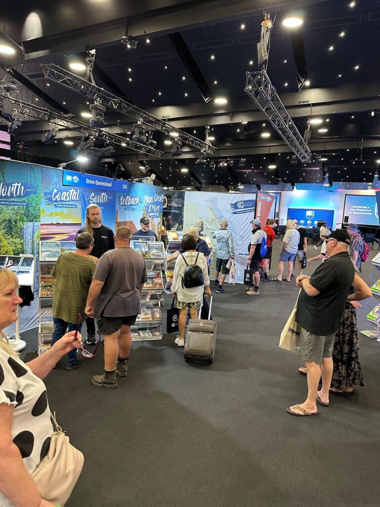 People stand and walk around an indoor exhibition with travel booths, informational displays, and promotional materials under bright lighting.