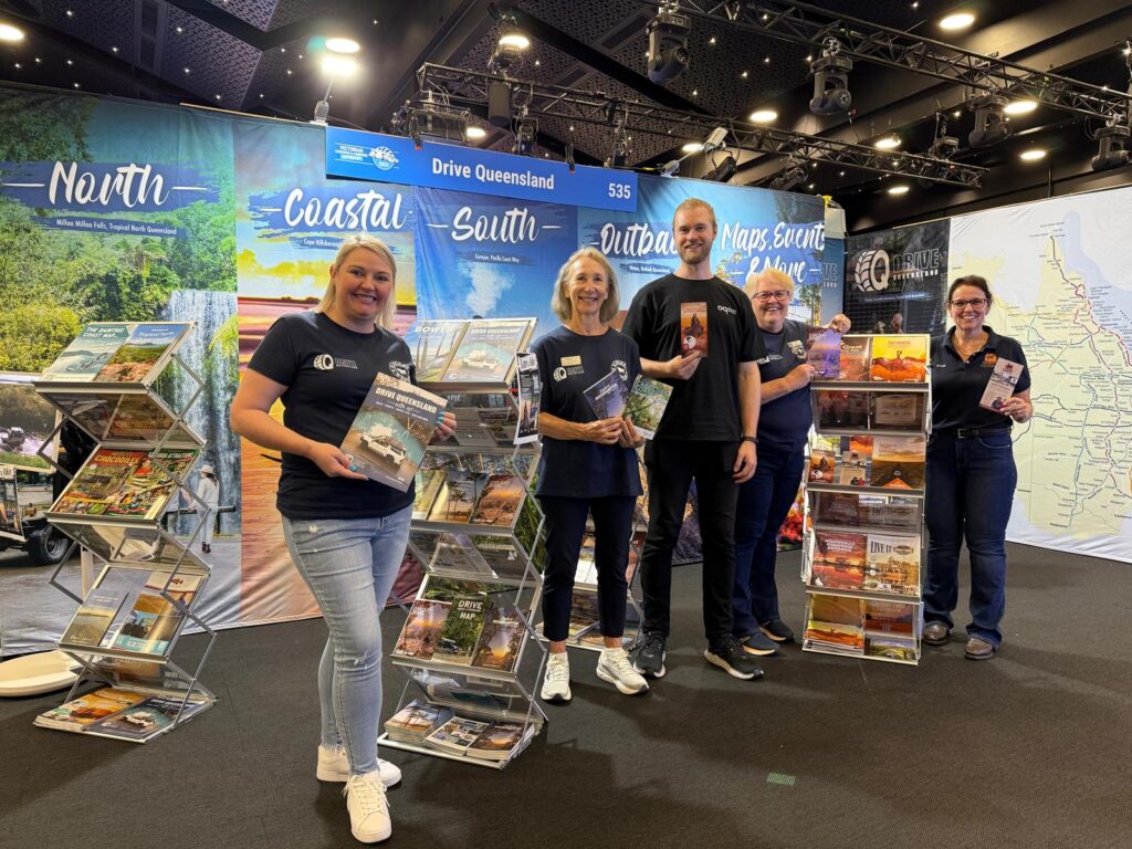 Five people stand at a tourism booth displaying Queensland travel brochures, with maps and promotional banners in the background.