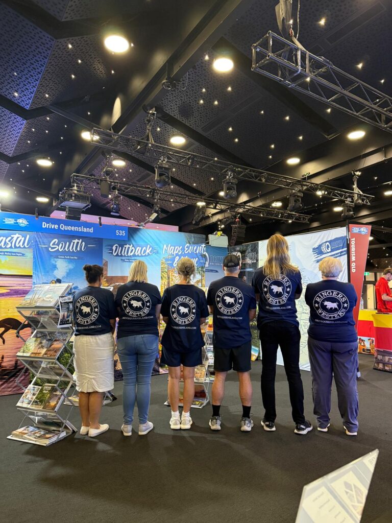 Six people stand with their backs to the camera at a tourism booth, wearing matching navy jackets with a circular logo, in an indoor exhibition space.