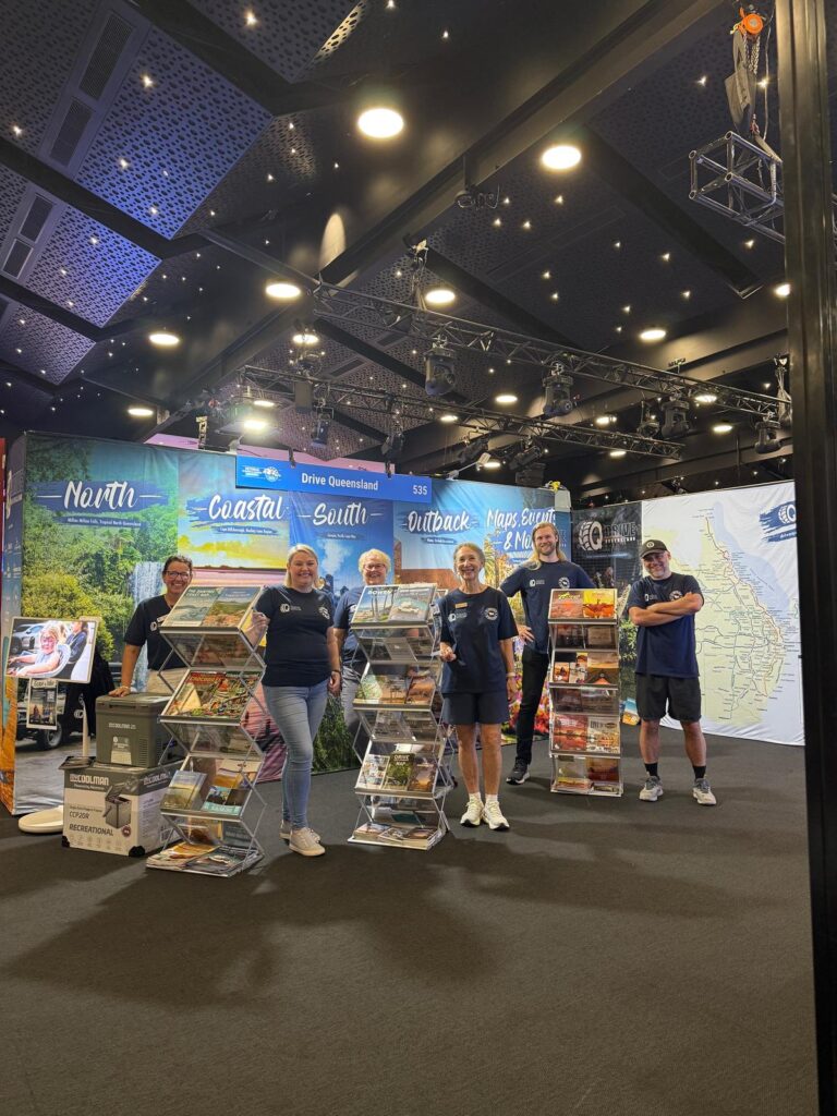 Six people stand at a tourism booth with brochures and regional banners under ceiling lights at an indoor event or expo.