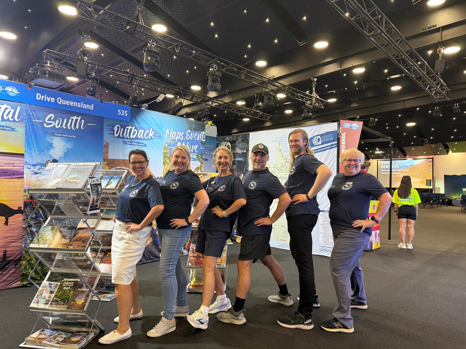 Six people in matching navy shirts pose side by side, smiling, at an indoor exhibition booth with travel brochures and banners promoting Queensland.