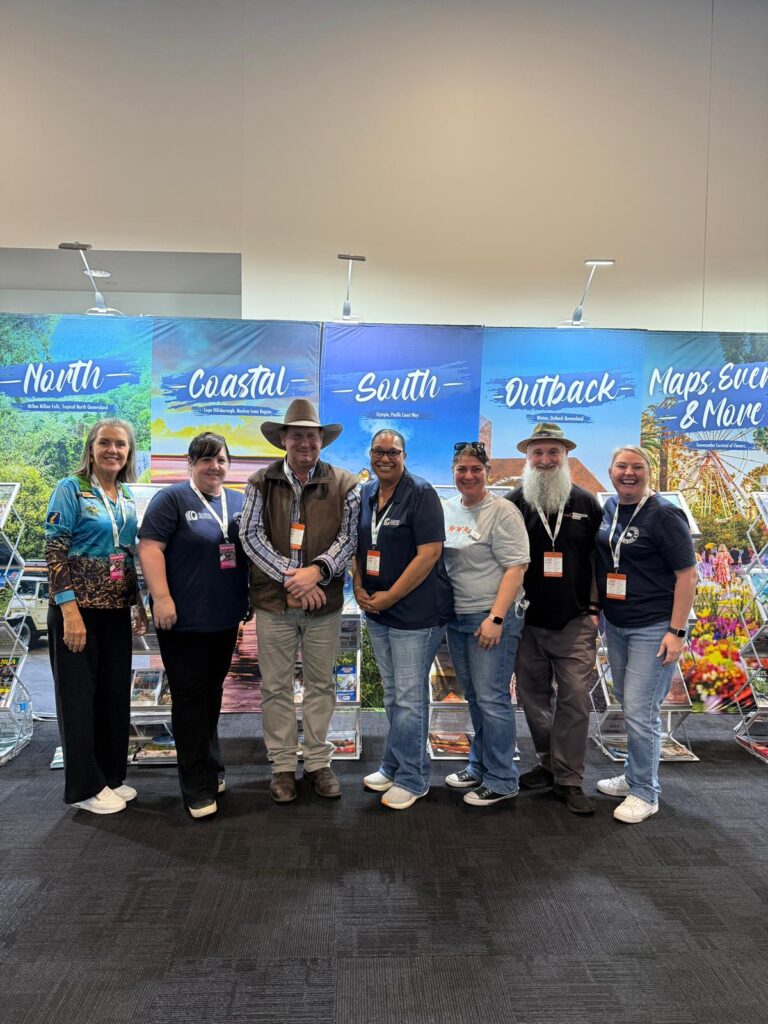 Seven people stand in a row posing for a group photo in front of a display with banners labeled North, Coastal, South, Outback, and Maps, Events & More.