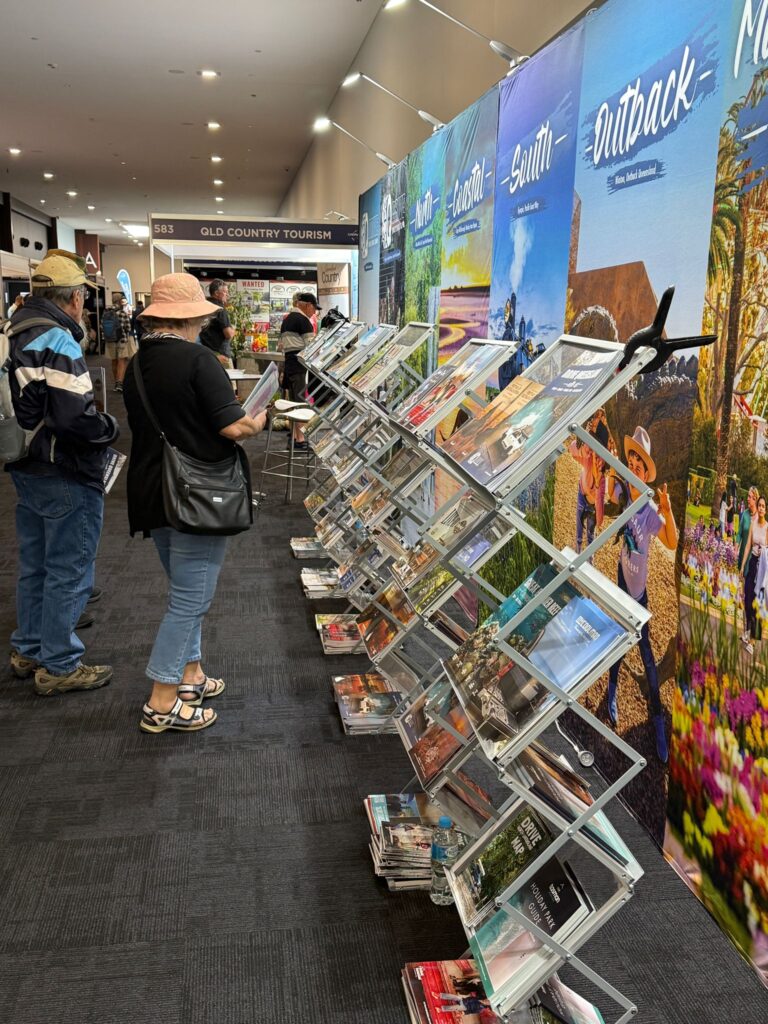 People stand and browse brochures on angled racks at a tourism expo, with colorful travel posters displayed on the wall behind them.