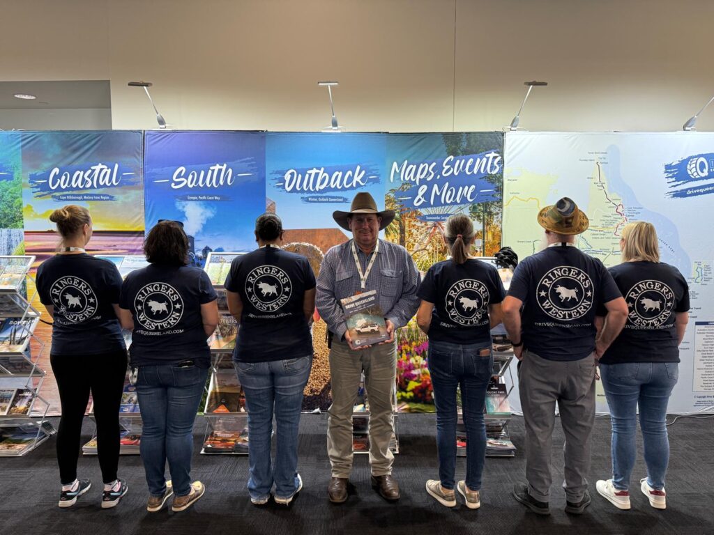 Seven people stand in front of a tourism display; six wear matching “Ringers Western” shirts, and one in a hat faces forward holding a brochure.