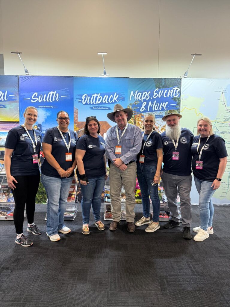 Seven people in matching navy shirts and event badges pose together in front of a booth with banners about maps, events, and the outback.