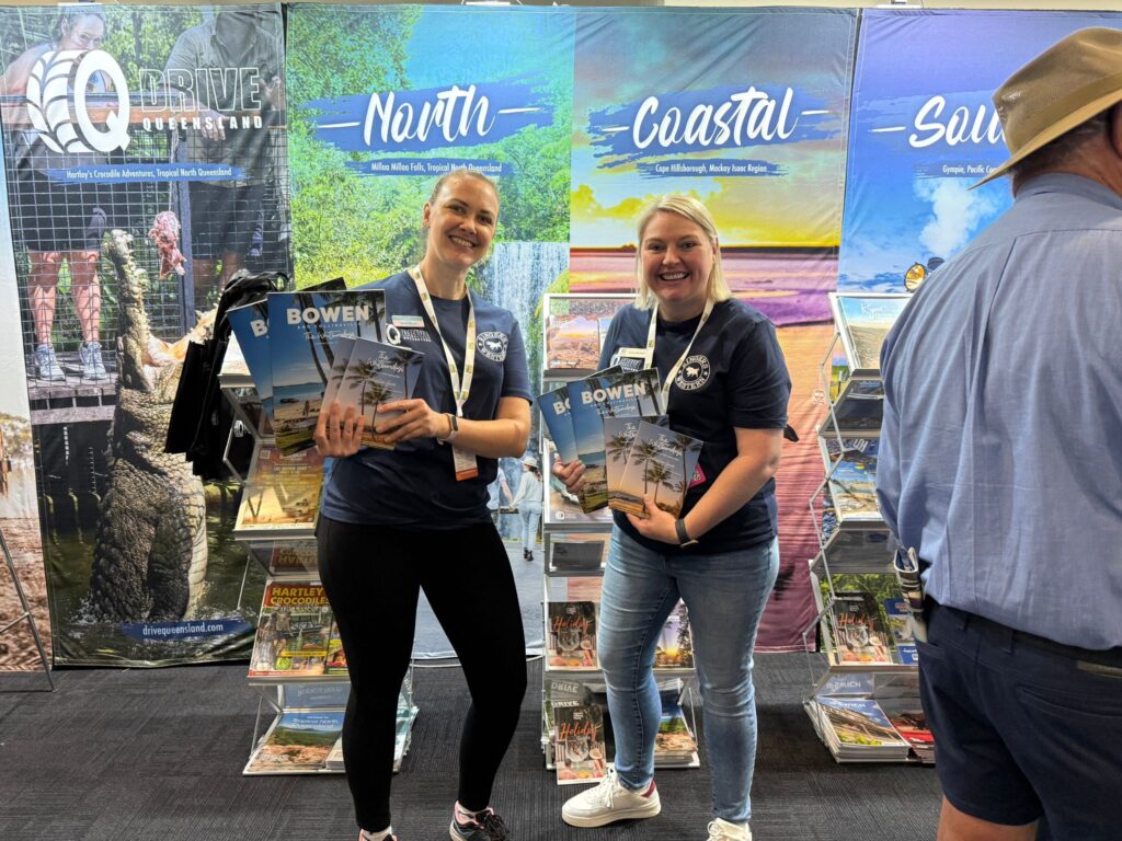 Two women stand in front of travel brochure displays, holding Bowen magazines and smiling at an indoor event; a man with a hat is partially visible on the right.