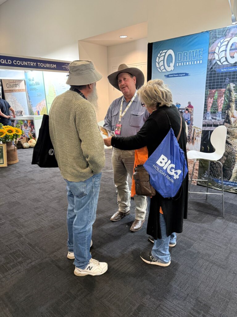 Three adults stand talking at a tourism expo; one man wears a wide-brim hat and badge, while the other two carry tote bags. Promotional banners and brochures are visible in the background.