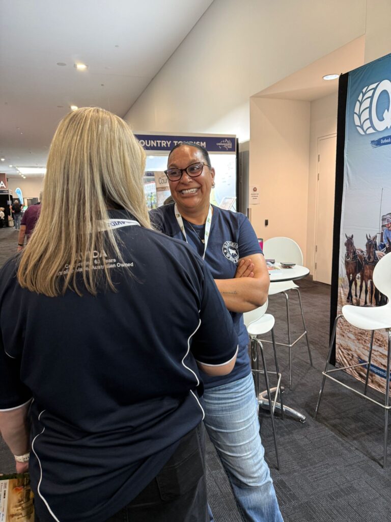 Two women in navy shirts converse at an indoor event; one faces the camera smiling with arms crossed, while the other has her back to the camera.