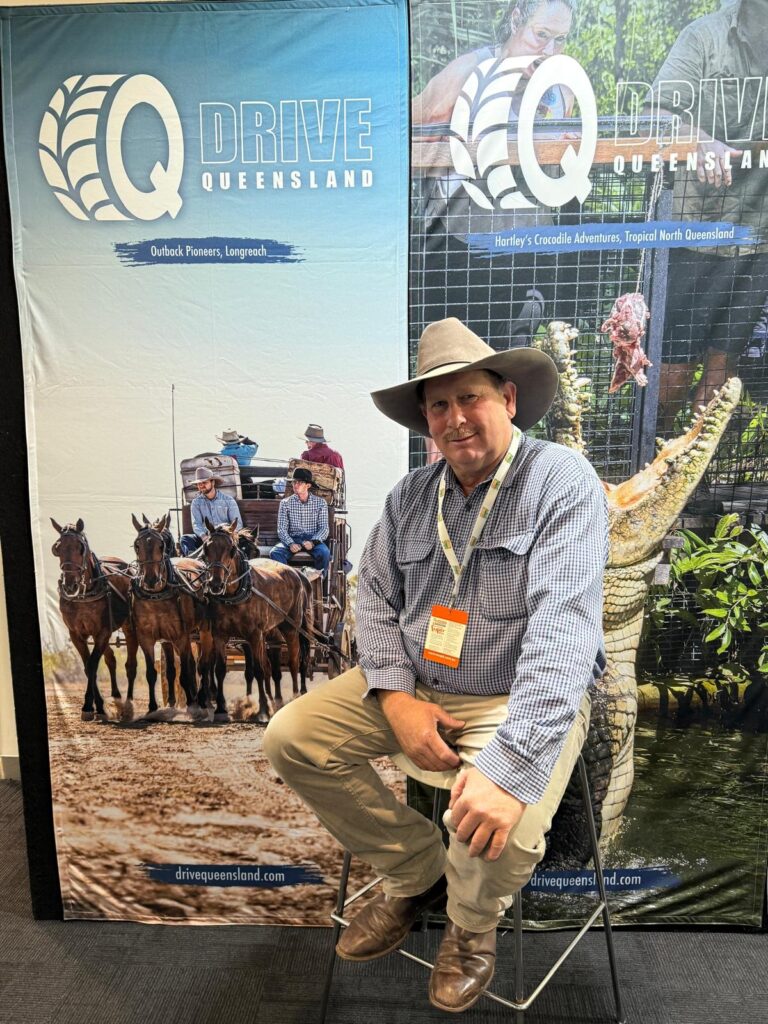 Man in a cowboy hat sits on a stool in front of banners promoting Queensland tourism, featuring images of horse-drawn wagons and a crocodile.