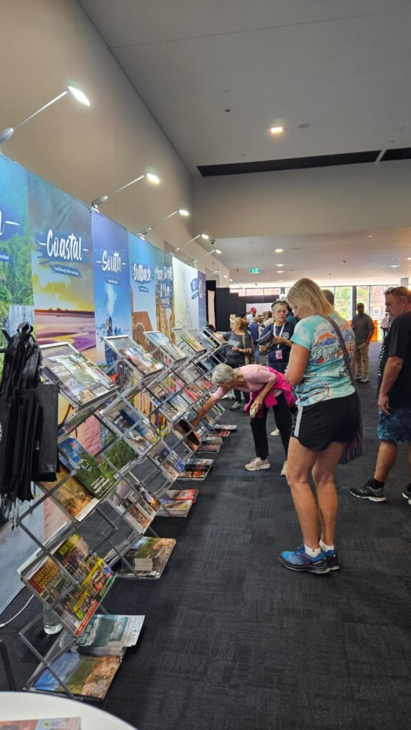 People browsing travel brochures displayed on racks at an indoor event with banners labeled 