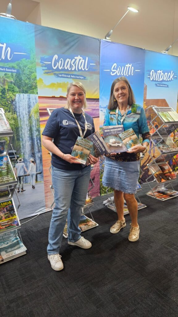 Two women stand indoors at a tourism booth, smiling and holding brochures, with display banners and racks of travel guides in the background.