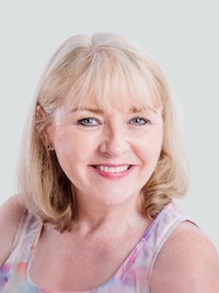 A middle-aged woman with blonde hair and light skin smiles at the camera against a plain light background. She is wearing a sleeveless, pastel-colored top.