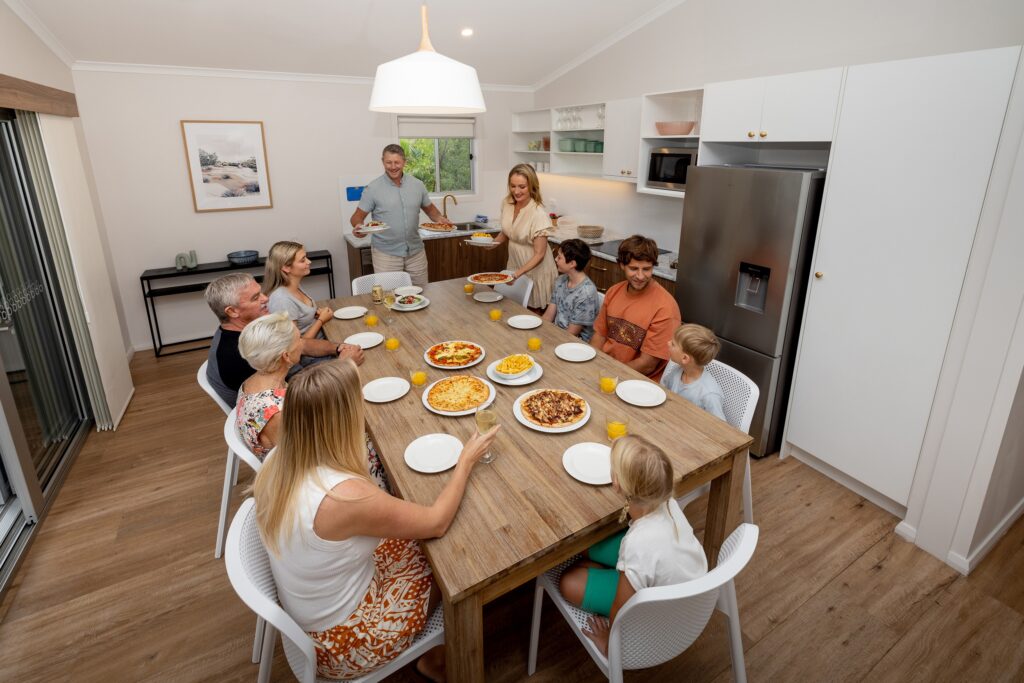 Eight people, including adults and children, sit around a wooden dining table set with pizza and orange drinks in a modern kitchen. Two adults are standing, serving food.