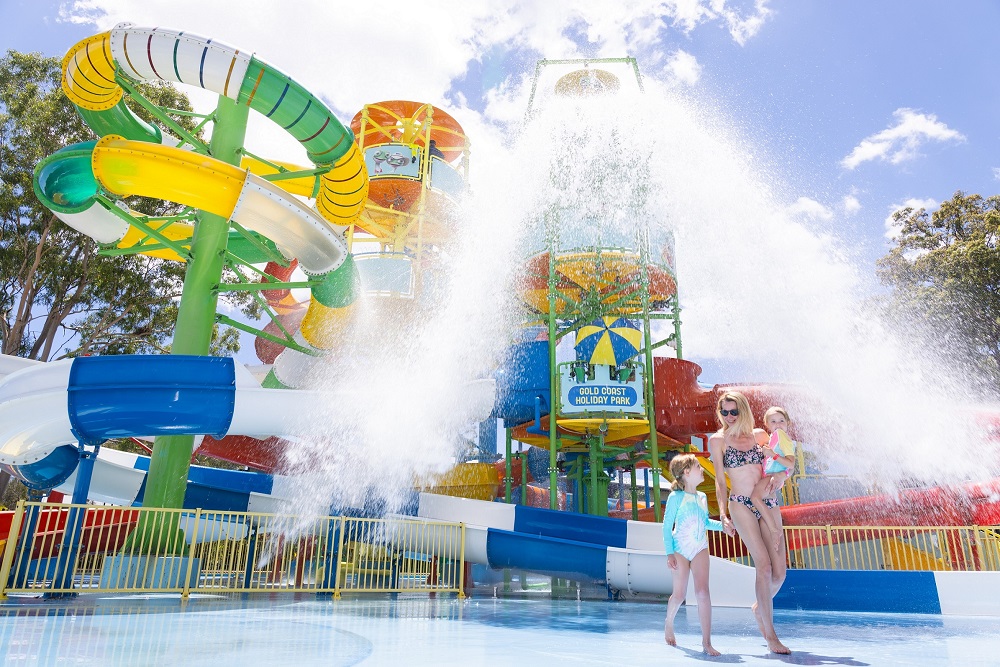 A woman and two children stand near colorful water slides as water sprays down at an outdoor water park on a sunny day.
