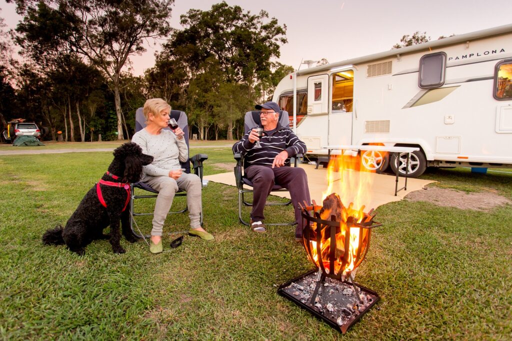 Two people sit in chairs by a campfire, holding drinks, with a black dog nearby and a caravan in the background on a grassy campsite.
