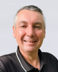 A middle-aged man with short gray hair smiles at the camera, wearing a black collared shirt with white trim, against a plain light background.