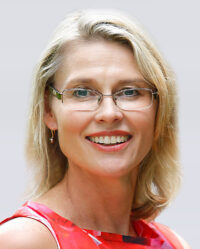 A woman with blonde hair, wearing glasses and a red patterned top, smiling at the camera against a plain light background.