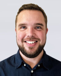A man with short brown hair, a beard, and a dark blue button-up shirt smiles at the camera against a plain light background.
