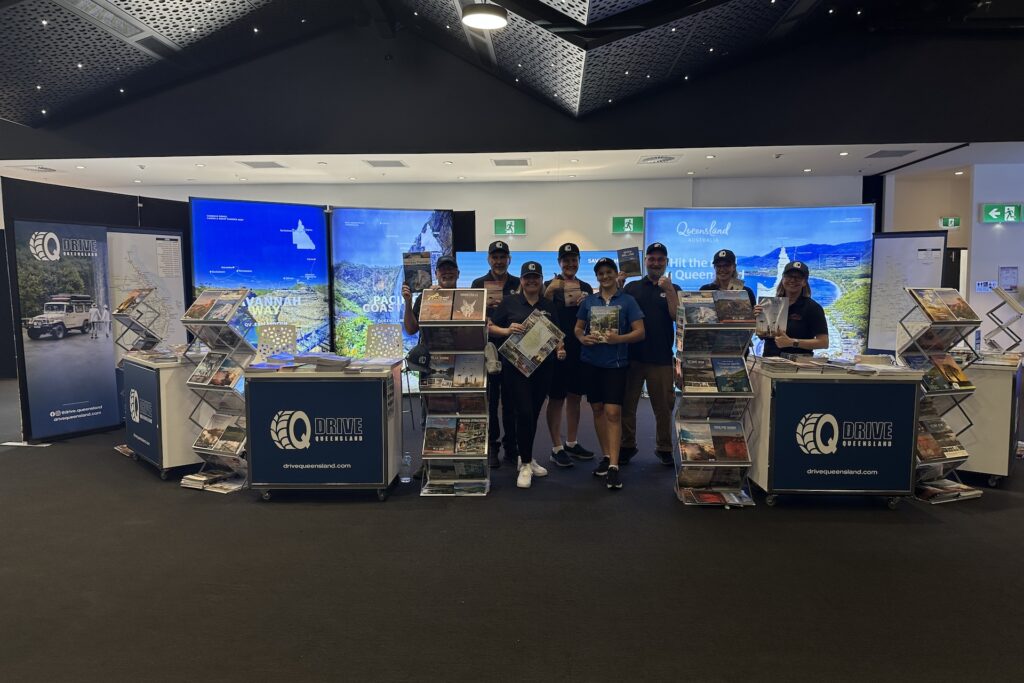 A group of people stands in front of a travel information booth with brochures and promotional banners in a convention center.
