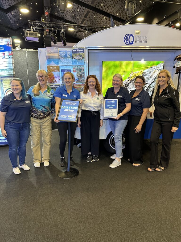 Seven women stand indoors next to an award sign and display, with two of them holding a plaque and certificate labeled 