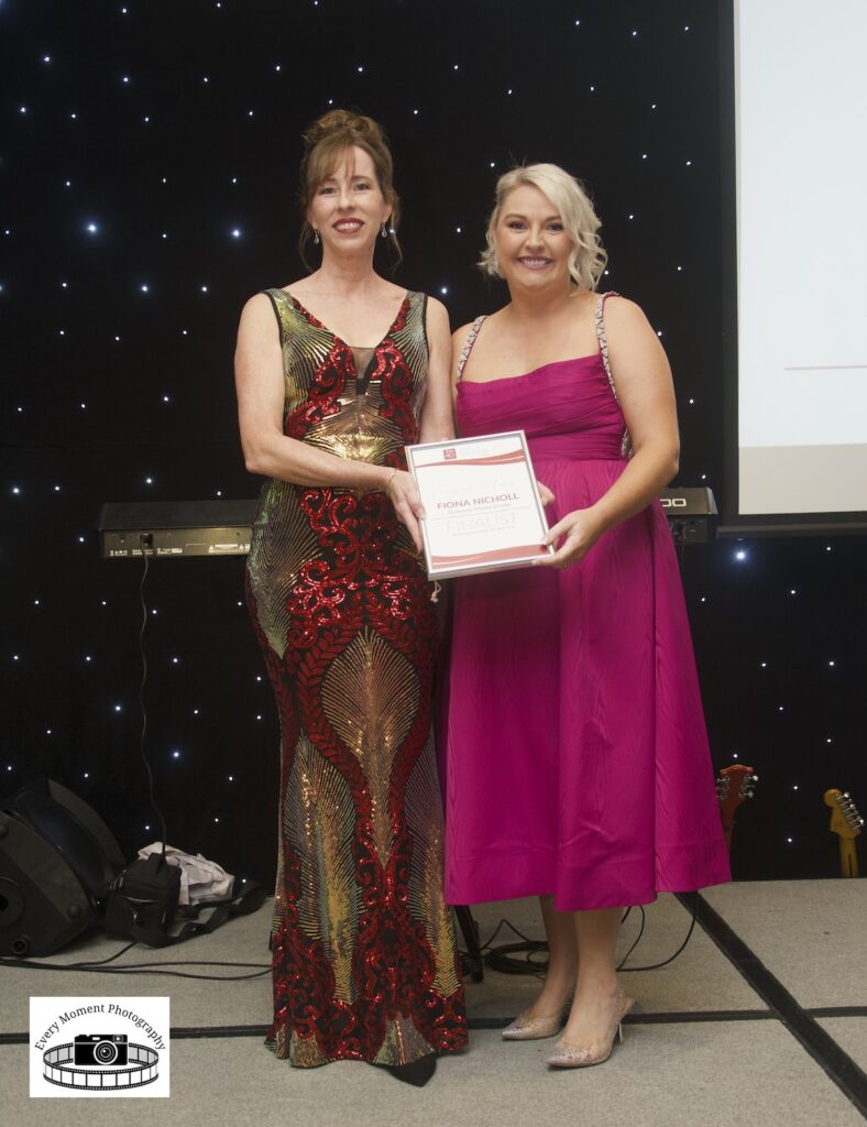 Two women in formal dresses stand on a stage. One holds a framed certificate. A starry backdrop and musical instruments are visible behind them.