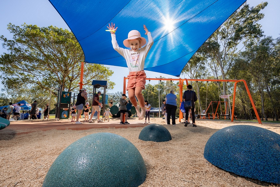 A young child in a pink hat jumps off a playground mound under a blue shade sail, with other children and adults in the background.