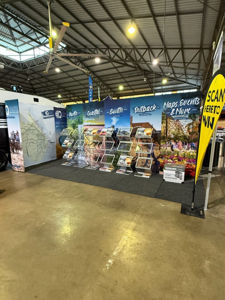 Tourism booth display with shelves of brochures and a backdrop labeled “North,” “Coastal,” “South,” “Outback,” “Maps, Events & More.” A yellow sign reads “SCAN HERE TO WIN.”.