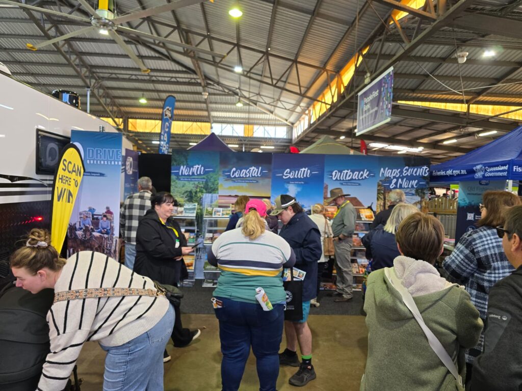 People are gathered at a travel expo booth with promotional banners, pamphlets, and a “Scan Here to Win” sign inside a large, well-lit exhibition hall.