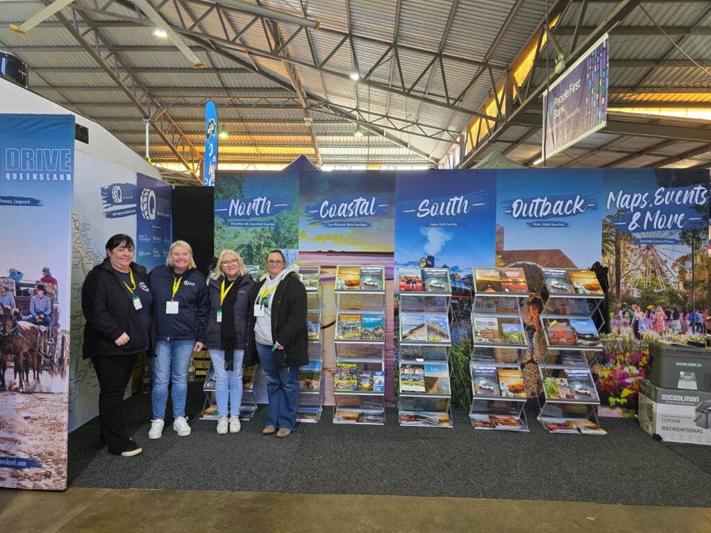 Four women stand in front of a tourism display booth featuring brochures and banners labeled North, Coastal, South, Outback, and Maps, Events & More in a large indoor venue.