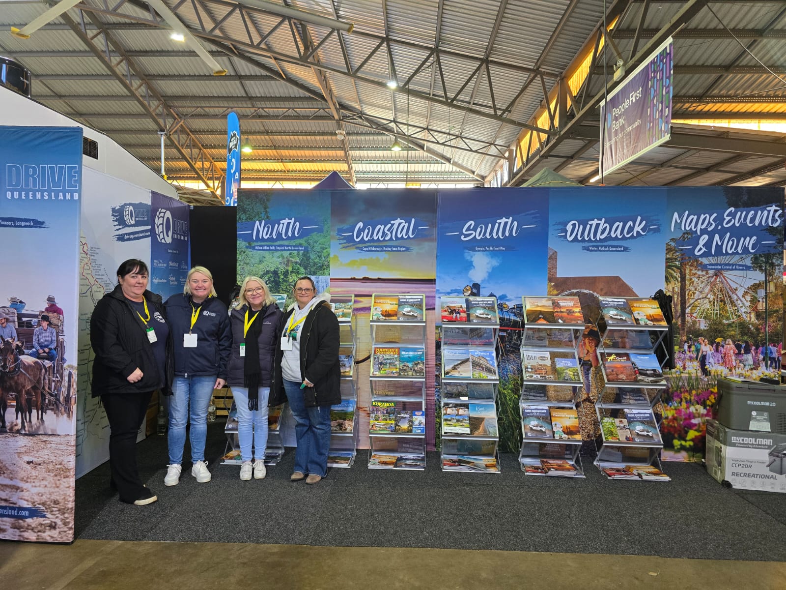 Four women stand in front of a tourism display booth featuring brochures and banners labeled North, Coastal, South, Outback, and Maps, Events & More in a large indoor venue.