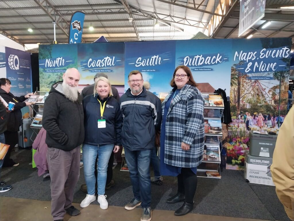 Four people stand smiling in front of a tourism booth with sections labeled North, Coastal, South, Outback, and Maps, Events & More inside an exhibition hall.