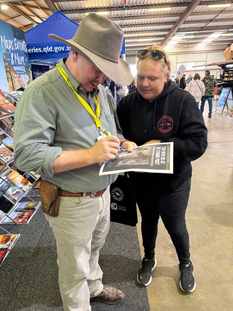 A man in a wide-brimmed hat signs a poster for a woman at an indoor event, with brochures and a blue information booth in the background.