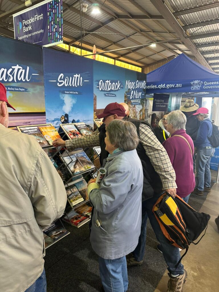 A group of people browse brochures at a tourism information stall featuring sections labeled Coastal, South, and Outback, inside an indoor event space.