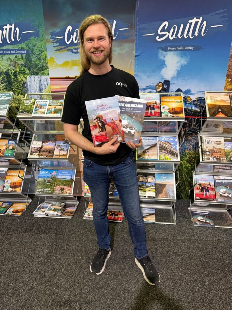 A person stands indoors in front of travel brochure displays, holding several tourism magazines and brochures.