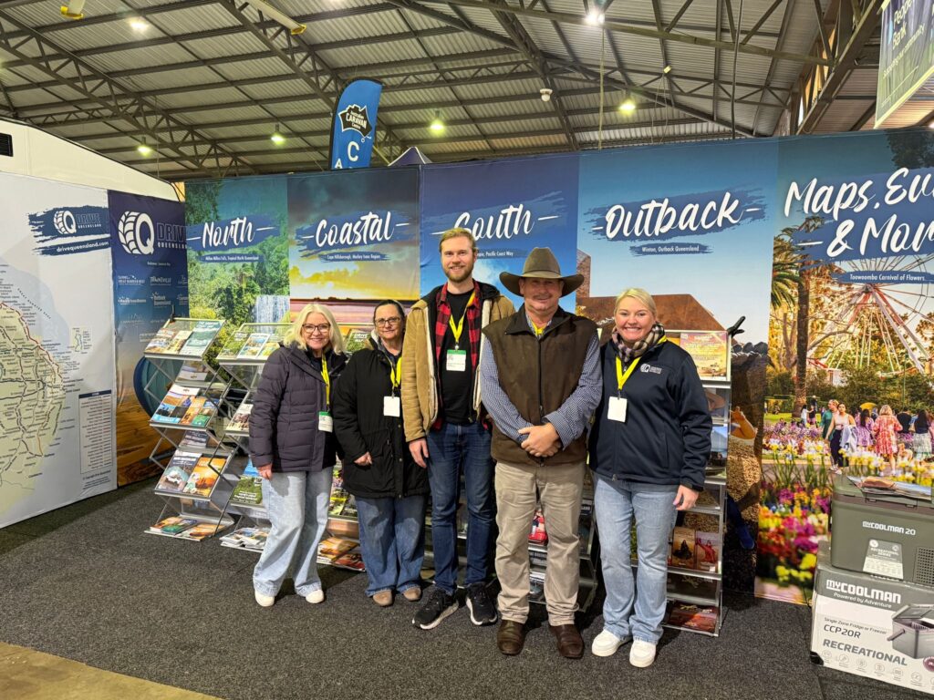 Five people stand and smile for a photo at an indoor tourism expo booth displaying brochures and large regional banners: North, Coastal, South, Outback, Maps, Events & More.