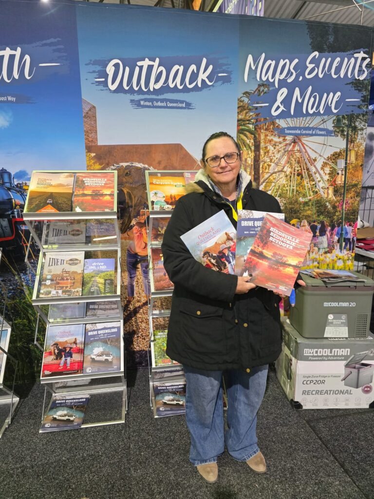 A woman in a black coat stands in front of a display of travel brochures labeled 