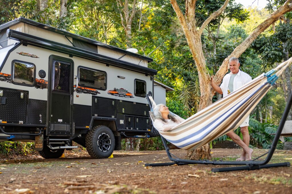 An older couple relaxes at a wooded campsite; one person lies in a hammock while the other stands nearby, next to a camper van.