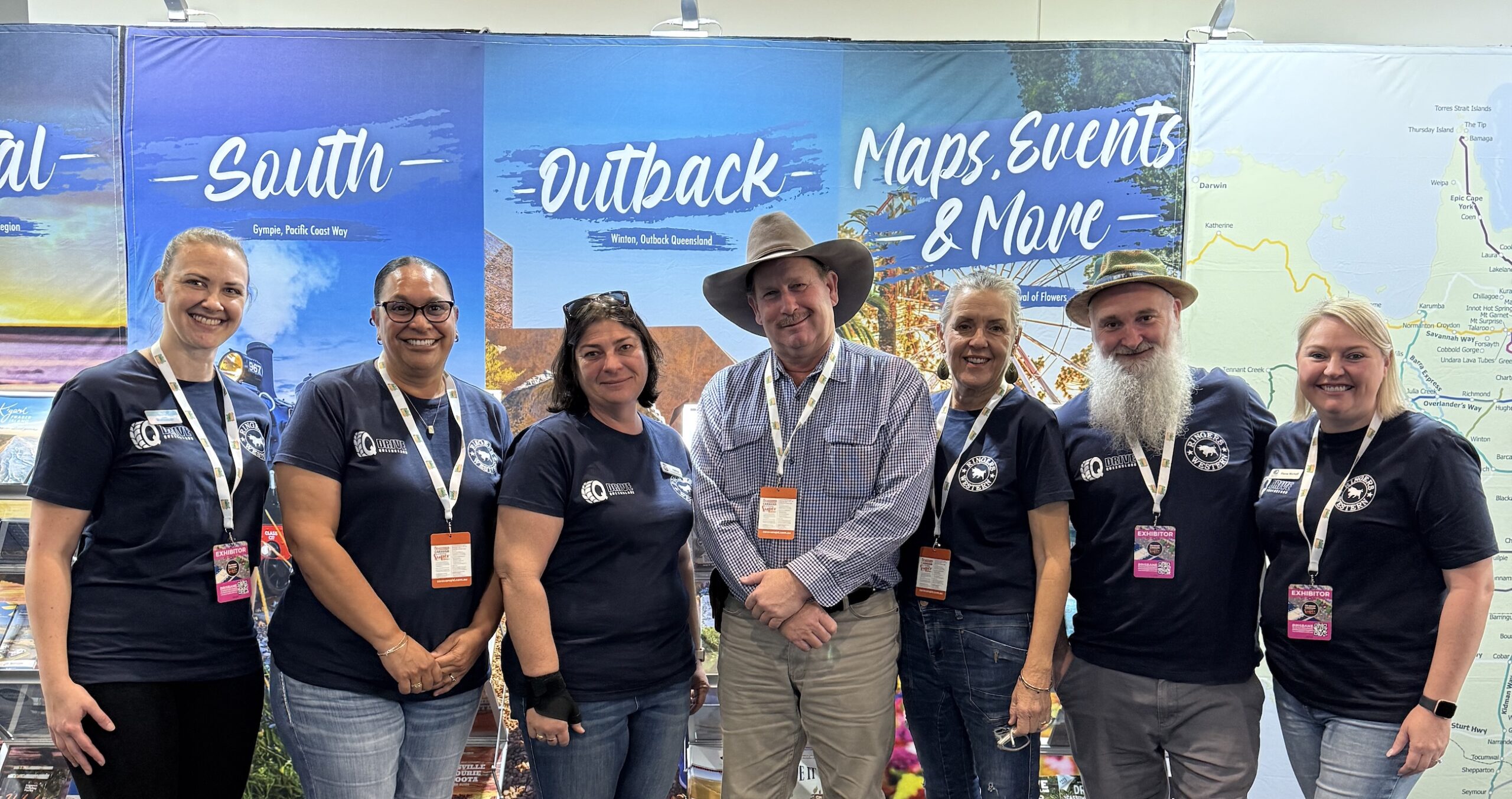 Seven people stand together indoors, smiling at the camera, wearing matching navy shirts and lanyards, in front of colorful travel-themed display banners.