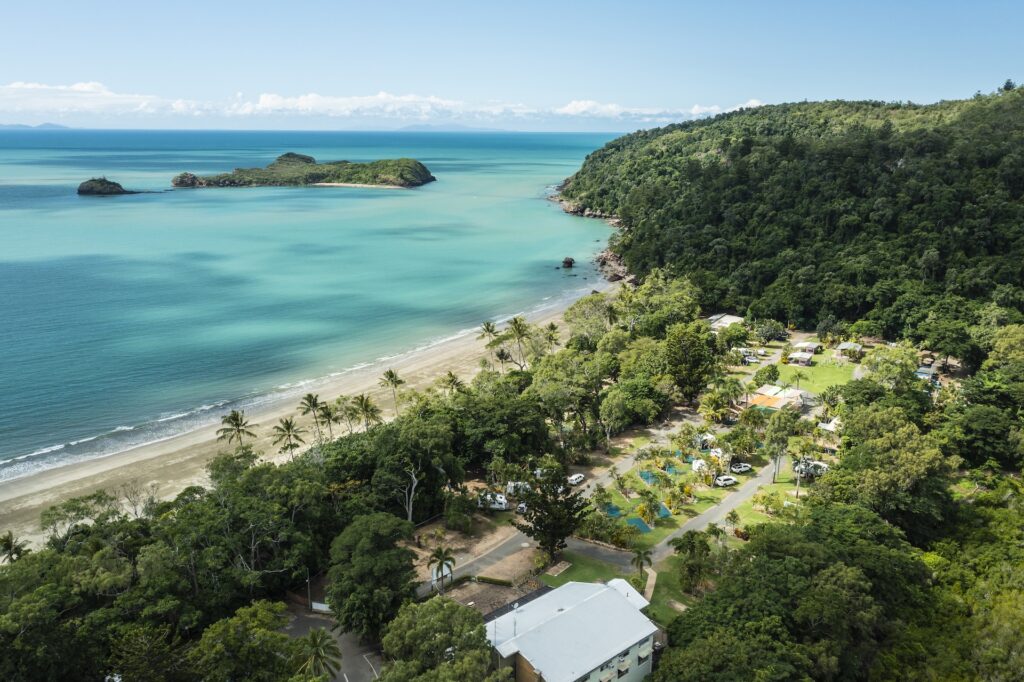 Aerial view of a coastal campsite with tents and cabins next to a sandy beach, turquoise water, and lush green forested hills.