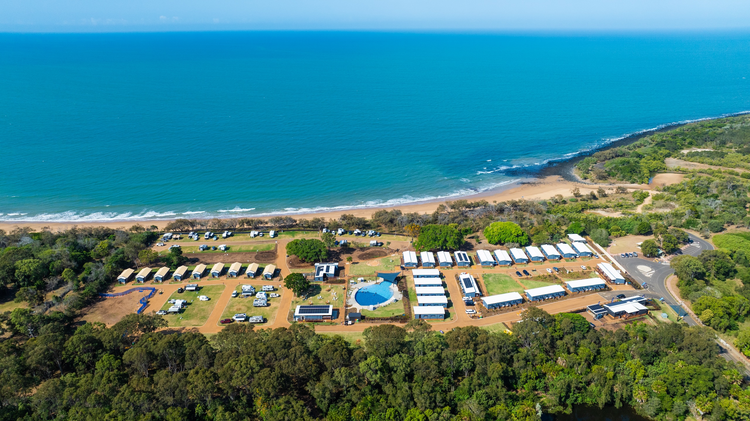Aerial view of a coastal campsite with cabins, tents, a swimming pool, and road access bordered by trees and a sandy beach along the blue ocean.