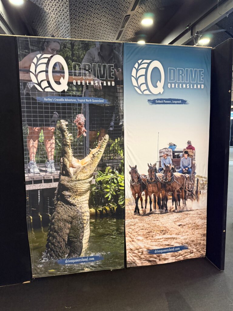 Two large banners display “Q Drive Queensland” with images of a jumping crocodile and people riding a horse-drawn carriage, advertising adventure tours in Queensland, Australia.