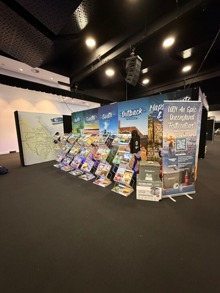 Tourism booth with maps, brochures, and promotional displays for Queensland travel, set up in an indoor event space with black flooring and ceiling.